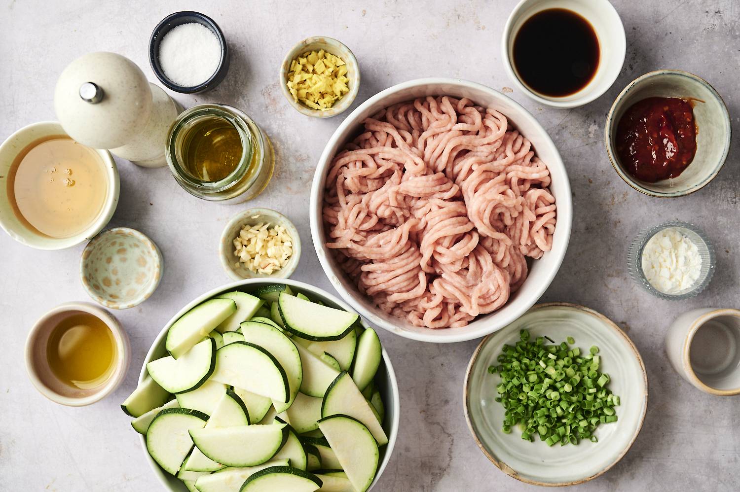 Overhead view of ground turkey, zucchini slices, green onions, garlic, ginger, soy sauce, chili paste, and other ingredients arranged in bowls and jars on a light surface for making ground turkey zucchini stir fry.