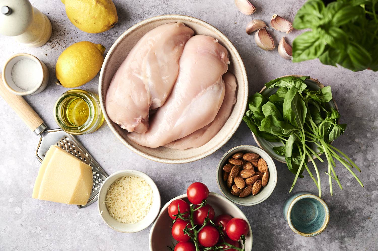 Fresh ingredients for pesto chicken laid out on a countertop, including raw chicken breasts, basil, cherry tomatoes, almonds, garlic, Parmesan, olive oil, and lemons.