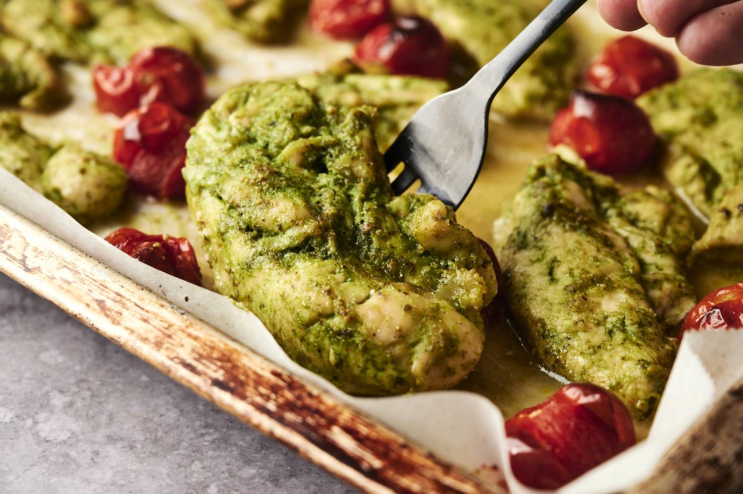 Close-up of baked chicken coated in vibrant green pesto with roasted cherry tomatoes on a parchment-lined baking sheet.