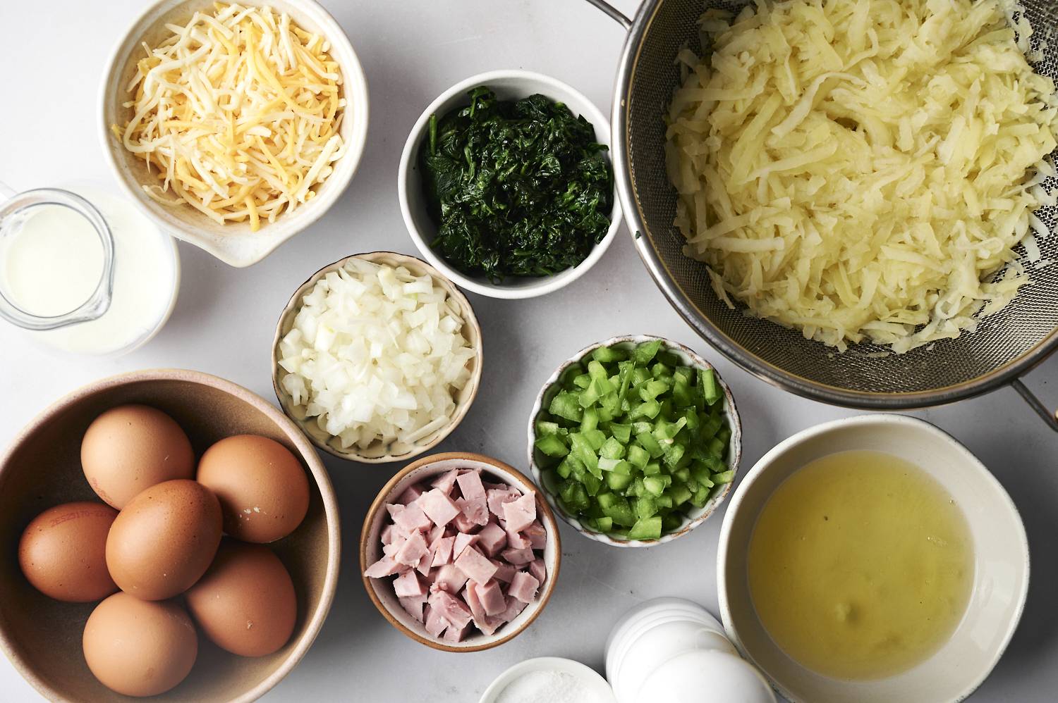 An overhead view of all the ingredients for a slow cooker breakfast casserole, including eggs, hash browns, cheese, spinach, onions, bell peppers, ham, and milk, arranged in small bowls.