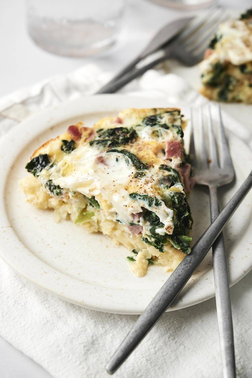 A close-up of a slice of breakfast casserole with layers of hash browns, eggs, ham, spinach, and cheese on a plate, with a fork resting beside it.