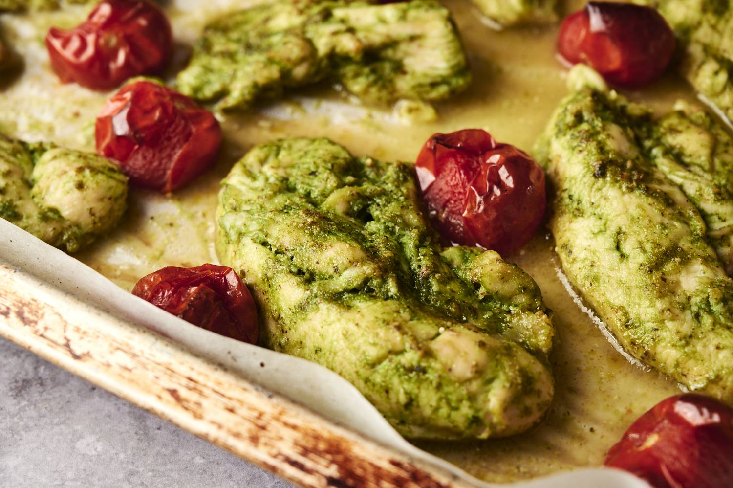 Close-up of juicy baked chicken coated in fresh pesto and surrounded by roasted cherry tomatoes on a parchment-lined baking sheet.