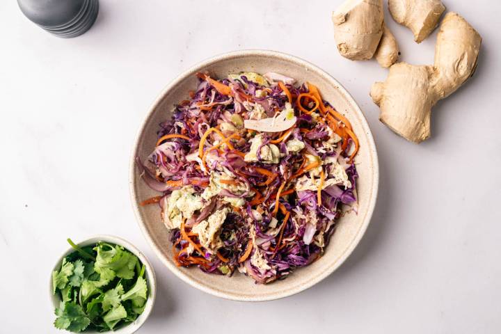 Cabbage Fried Rice A colorful salad with shredded purple cabbage, carrots, and dressing in a beige bowl on a white surface. Fresh cilantro and ginger root surround it, suggesting fresh ingredients.