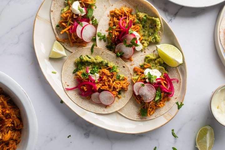 Cochinita Pibil Cochinita pibil tacos with marinated shredded pork, guacamole, cilantro, pickled red onions, and limes in corn tortillas.