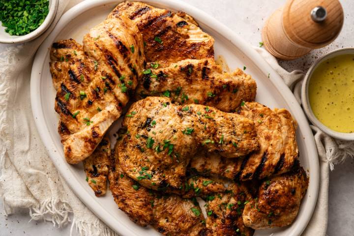 Copycat Chick-fil-A Grilled Chicken Grilled chicken breasts garnished with chopped parsley on a white plate. A small bowl of green herbs and a bowl of yellow sauce are nearby.