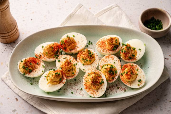Easy Healthy Deviled Eggs A plate of deviled eggs garnished with paprika and chives is set on a beige napkin. A small bowl of chives and a pepper mill are nearby.