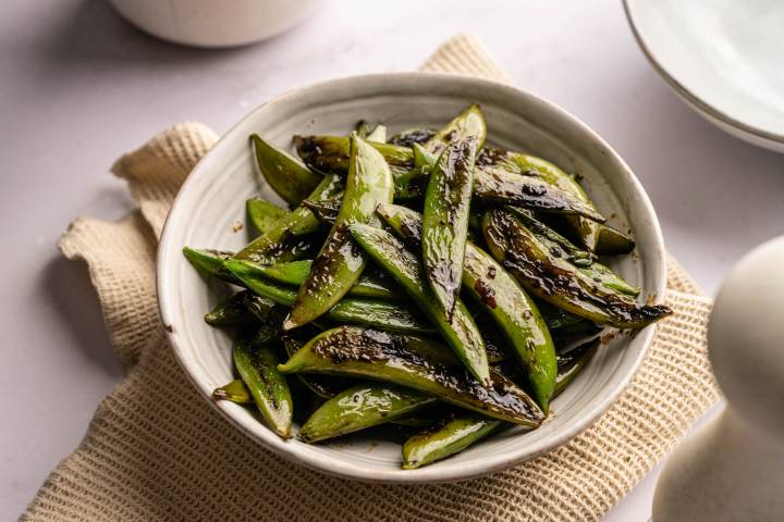 Garlic Sugar Snap Peas A white bowl filled with glossy, sautéed sugar snap peas on a textured beige cloth.