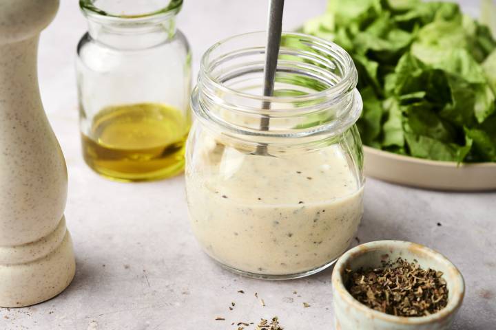 Greek Salad Dressing Jar of creamy Greek salad dressing with herbs, olive oil, and fresh lettuce in the background.