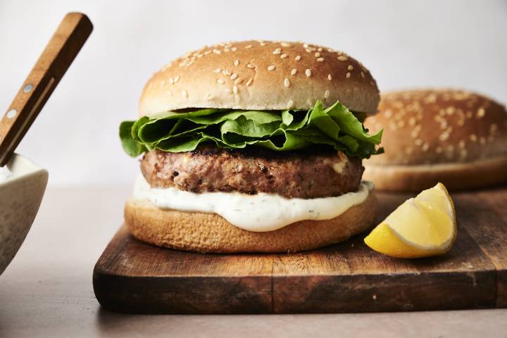 Greek Turkey Burgers Greek turkey burger on a wooden board with crisp lettuce and creamy tzatziki sauce inside a sesame seed bun, garnished with a lemon wedge on the side.