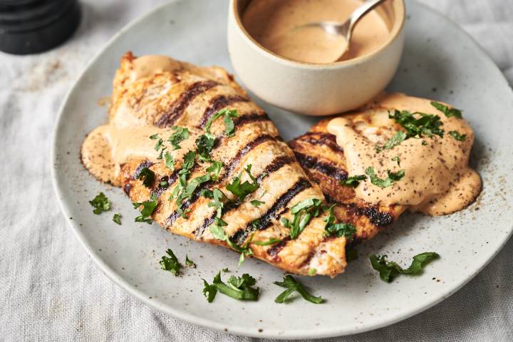 Grilled Chipotle Ranch Chicken Grilled chicken breasts with char marks topped with creamy sauce and cilantro on a plate, next to a bowl of sauce with a spoon, set on a light tablecloth.
