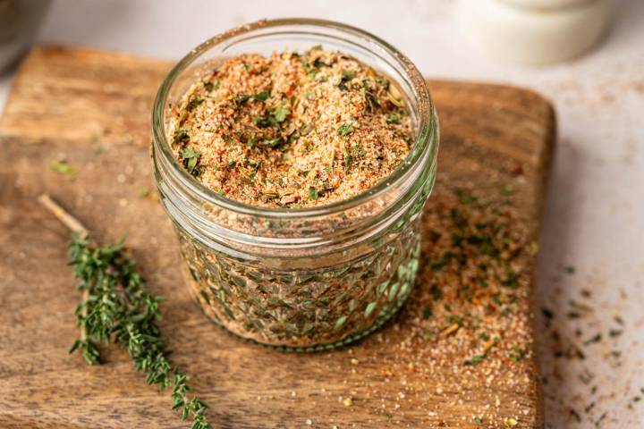 Homemade Italian Sausage Seasoning A glass jar filled with a colorful seasoning blend sits on a wooden board. Fresh thyme sprigs are nearby, highlighting a rustic kitchen setting.