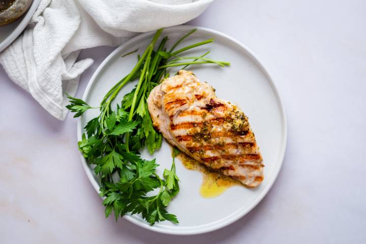 Lemon Pepper Marinade Grilled chicken breast with herb seasoning, served on a white plate with fresh parsley. Simple and appetizing presentation on a light background.