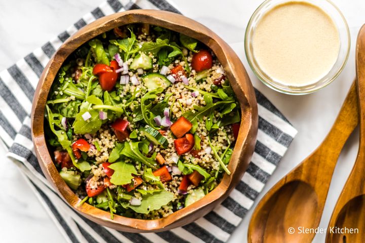 Mediterranean Quinoa Bowl Mediterranean quinoa bowl with quinoa, arugula, red peppers, tomatoes, and hummus dressing in a wooden bowl.