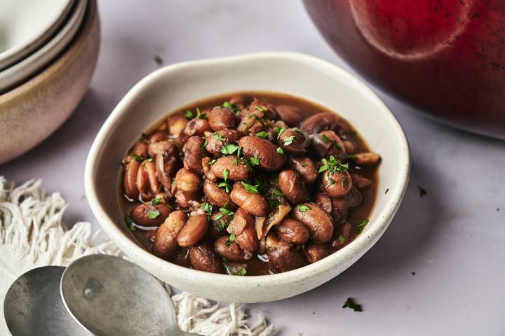How to Cook Beans in the Oven A bowl of savory, cooked beans garnished with fresh parsley, accompanied by shiny metal spoons and delicate dishware, set on a table.