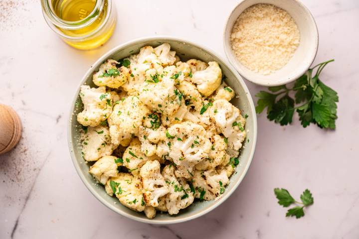 Sautéed Cauliflower A bowl of roasted cauliflower florets garnished with chopped parsley on a marble surface. Nearby are a bowl of grated cheese and a jar of olive oil.