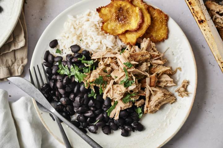 Slow Cooker Garlic Cuban Pork Shredded garlic Cuban pork served with black beans, rice, and crispy plantains on a white plate. The pork is topped with fresh cilantro, and there is a fork and knife on the side.
