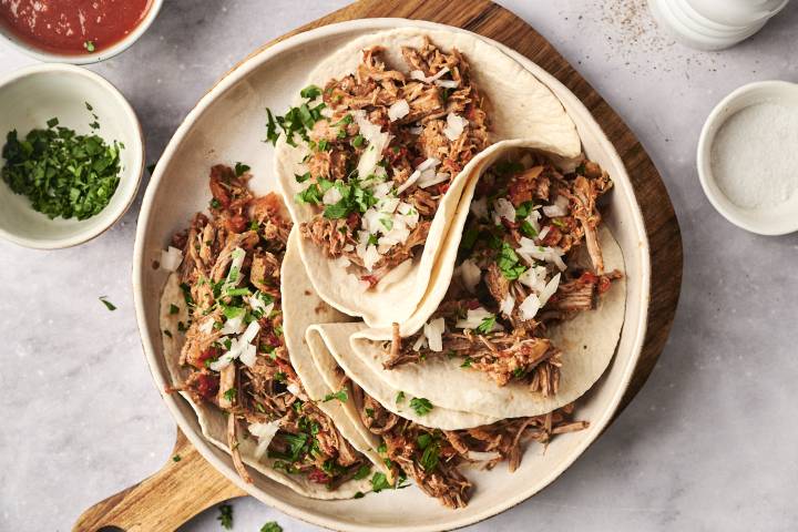 Slow Cooker Spicy Braised Beef A plate of soft flour tortillas filled with slow-cooked spicy braised beef, topped with chopped onions and fresh cilantro, served with salsa on the side.