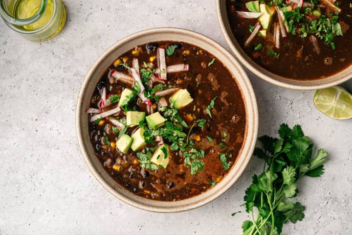 Spicy Black Bean and Corn Soup A bowl of black bean soup topped with avocado, cilantro, and radish strips on a textured gray surface. A jar of oil and fresh lime and cilantro are nearby.