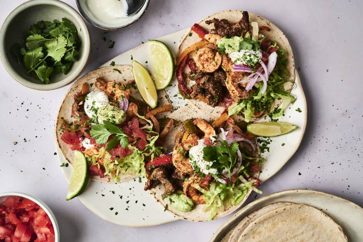 Steak and Shrimp Fajitas Three vibrant tacos topped with shrimp, beef, lettuce, tomatoes, onions, and cilantro, served with lime wedges and dipping sauce.