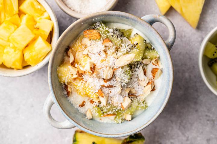 Tropical Yogurt Bowl A bowl of yogurt topped with chopped almonds, shredded coconut, kiwi slices, and pineapple chunks. Surrounding it are bowls of pineapple and kiwi on a gray surface.