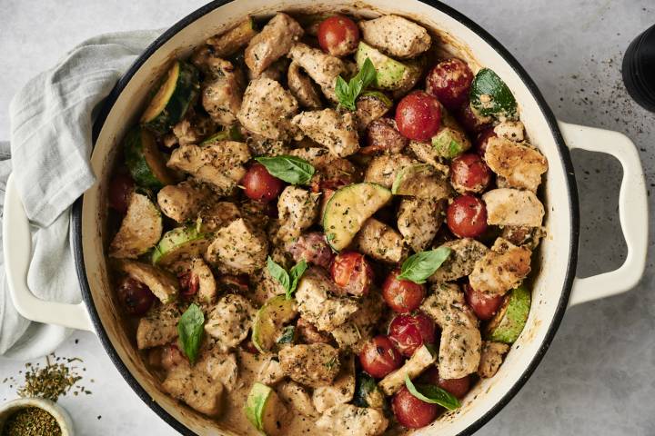Healthy Tuscan Chicken {Olive Garden Copycat) Top-down view of a creamy Tuscan chicken skillet filled with chunks of seasoned chicken, cherry tomatoes, zucchini slices, and fresh basil in a creamy herb sauce. A gray napkin and small bowl of Italian seasoning rest nearby.