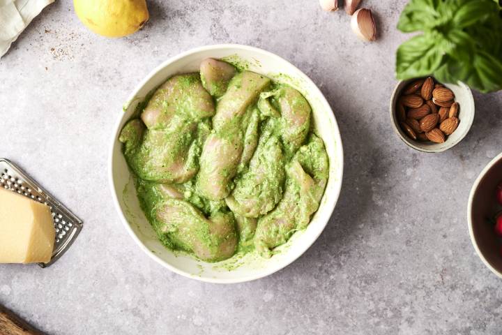 Pesto Chicken Raw chicken breasts coated in vibrant green pesto marinade, ready to bake, surrounded by fresh ingredients like basil, garlic, almonds, and Parmesan.