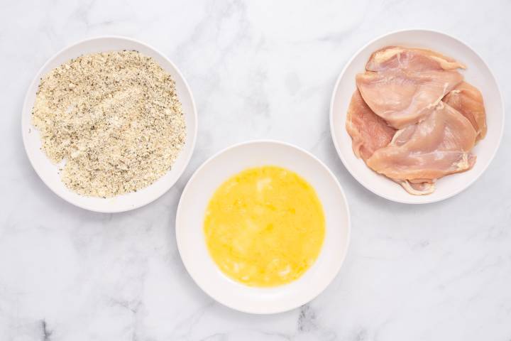 Sheet Pan Healthy Chicken Parm Bread crumbs, eggs, and chicken breast on a marble board.