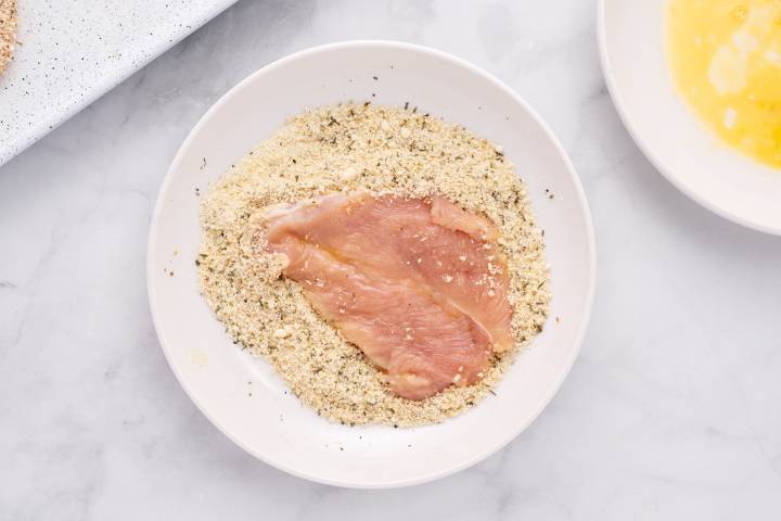 Sheet Pan Healthy Chicken Parm Chicken breast being dredged in bread crumbs and Parmesan cheese.