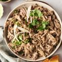 Easy Healthy Carnitas - Slow Cooker or Instant Pot A plate of shredded pork carnitas garnished with sliced onions, green onions, and cilantro, served with tortillas and dipping sauce on the side.