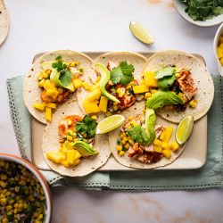 Salmon Tacos with Corn Salsa Salmon tacos with corn salsa, mango, fresh avocado slices, and lime wedges on a tray.