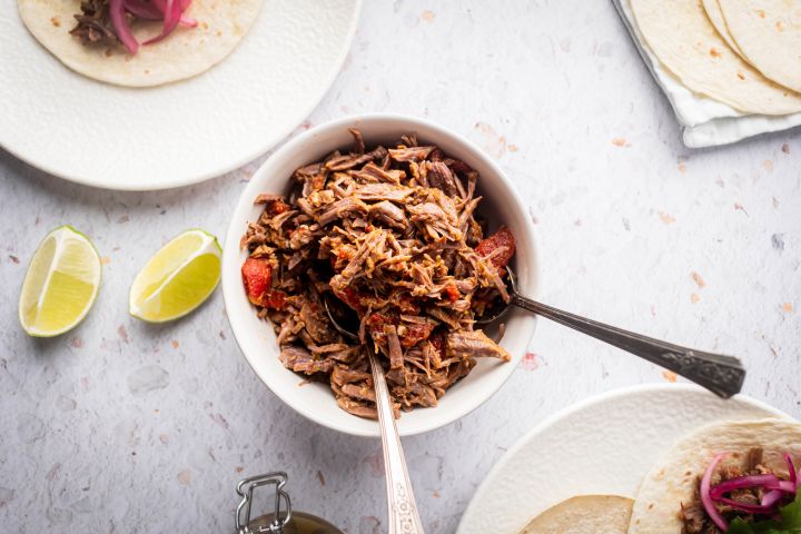 Slow Cooker Mexican Shredded Beef Mexican shredded beef in a bowl with tomatoes and chipotle peppers with tacos on the side.