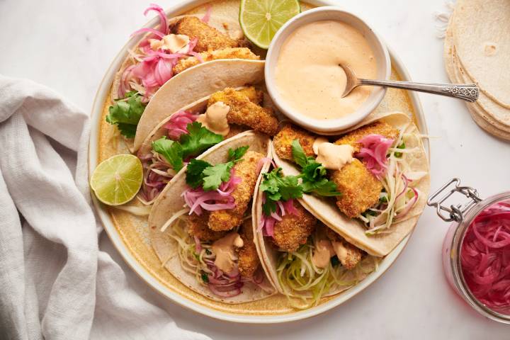 Baja Fish Tacos Baja fish tacos with crispy battered fish, cabbage slaw, pickled red onions, and white sauce served in corn tortillas on a plate.