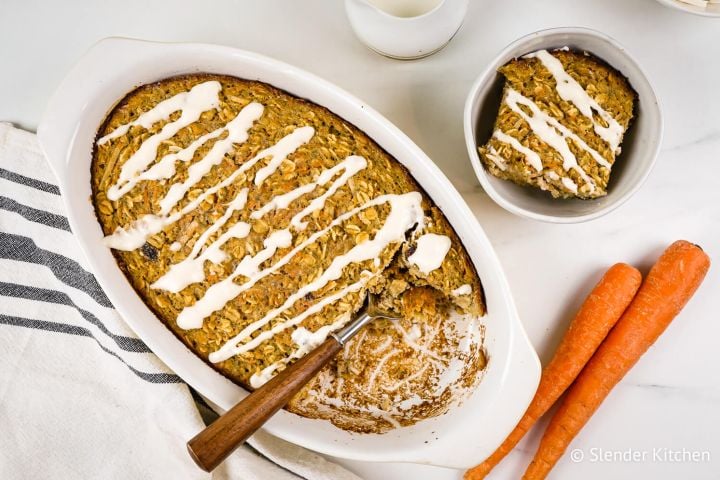 Baked Carrot Cake Oatmeal with Cream Cheese Glaze Baked carrot cake oatmeal with a cream cheese glaze in a baking dish with shredded coconut and oats.