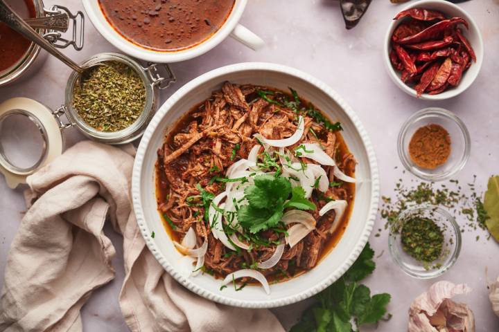 Beef Birria (Slow Cooker, Instant Pot, or Dutch Oven) Birria de res with a red chile sauce served in a bowl with corn tortillas, sliced onions, cilantro, and consome.