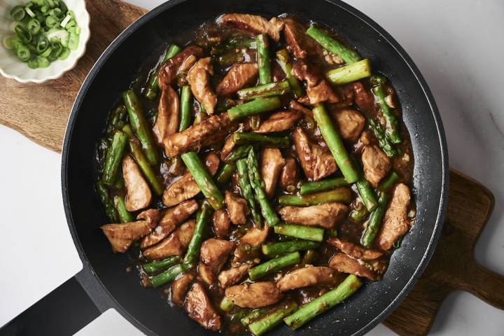 Chicken and Asparagus Stir Fry Stir-fried chicken and asparagus in a dark sauce, garnished with chopped green onions in a bowl on the side.