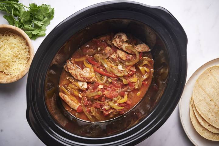 Slow Cooker Chicken Fajitas (6 Ingredients) Slow cooker chicken fajitas with bell peppers, onions, sliced chicken breast, and tomatoes in a black slow crockpot with tortillas on the side.