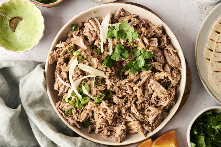 Easy Healthy Carnitas - Slow Cooker or Instant Pot A plate of shredded pork carnitas garnished with sliced onions, green onions, and cilantro, served with tortillas and dipping sauce on the side.