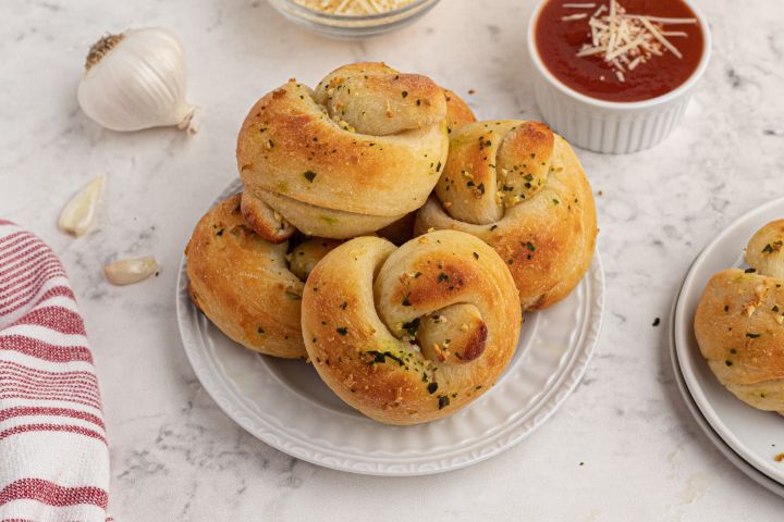 Garlic Knots (with Two Ingredient Dough) Garlic knots made with two ingredient dough topped with fresh garlic, melted butter, and basil on a plate with marinara sauce on the side.