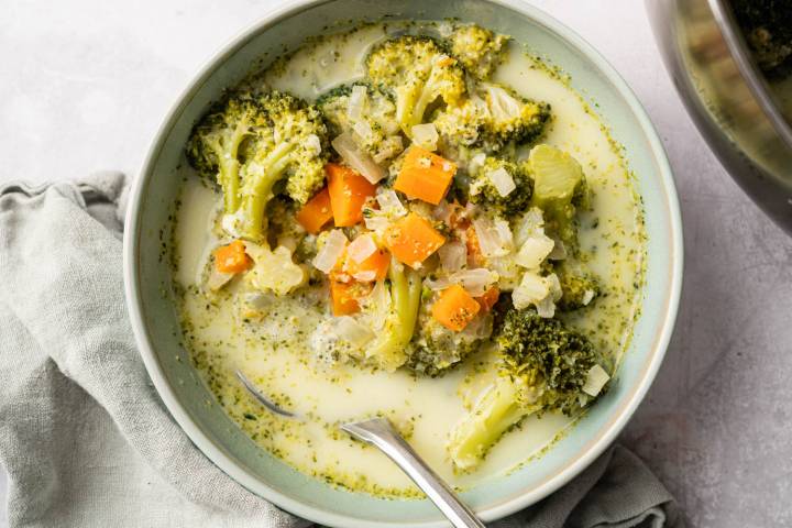 Healthy Broccoli Cheese Soup A bowl of broccoli cheddar soup with visible broccoli florets, diced carrots, and onion chunks in a creamy broth. A spoon rests on the bowl's edge. The soup sits on a light-colored cloth, conveying a comforting, homemade feel.