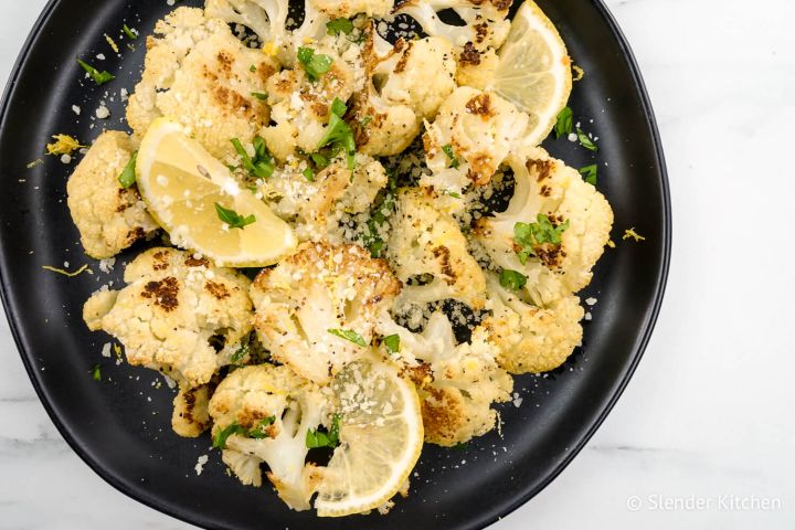 Lemon Garlic Roasted Cauliflower Lemon garlic roasted cauliflower with Parmesan cheese and lemon slices on a black plate.