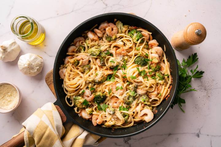 Lemon and Broccoli Pasta Shrimp A skillet of shrimp and broccoli linguine, garnished with parsley and Parmesan. Surrounding are garlic cloves, olive oil, and a pepper mill on a marble surface.