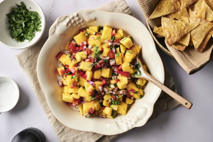 Mango Salsa Fresh mango salsa in a cream-colored bowl with a wooden spoon, made with diced mango, red onion, red bell pepper, jalapeño, and cilantro, served with tortilla chips on the side.