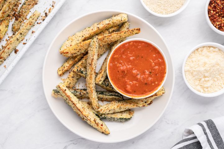 Oven Baked Parmesan Zucchini Fries Baked Parmesan zucchini fries with a crispy panko breading on a white plate with marinara sauce.