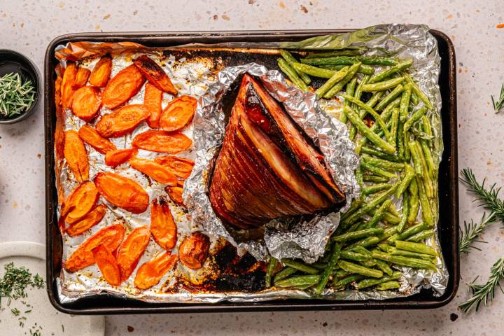 Sheet Pan Easter Ham and Vegetables A roasted ham surrounded by glazed carrots and green beans on a foil-lined baking sheet.