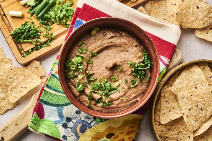 Shortcut Refried Beans A bowl of creamy refried beans is garnished with chopped green onions and cilantro, served alongside tortilla chips on a colorful napkin.