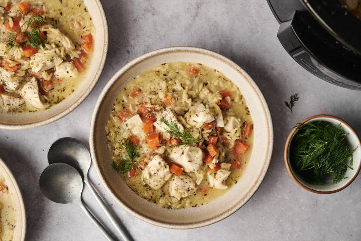 Slow Cooker Greek Lemon Chicken Orzo Soup Two bowls of creamy chicken and rice soup garnished with fresh dill and diced tomatoes, accompanied by two spoons and a bowl of herbs.