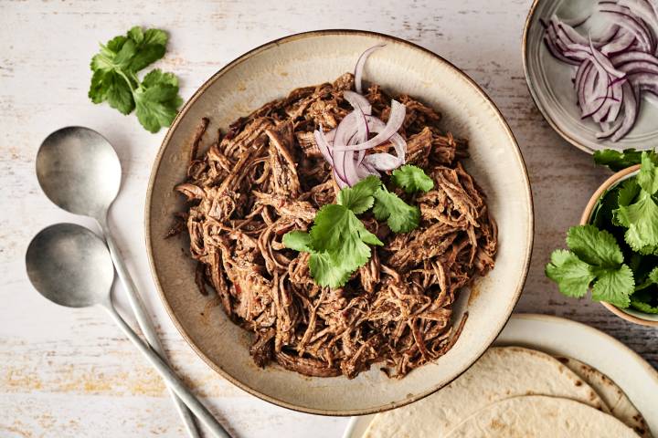 Slow Cooker Mexican Beef Adobo A bowl of tender, shredded Mexican beef adobo is garnished with fresh cilantro and sliced red onions, ready to be served with tortillas.