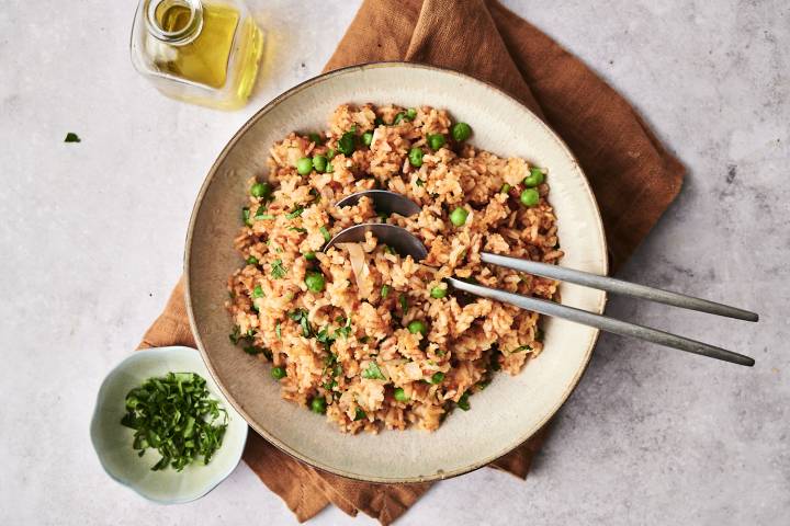 Slow Cooker Mexican Rice Overhead view of a bowl of slow cooker Mexican rice with green peas and chopped cilantro, served with two metal spoons on a brown napkin, next to a small bowl of herbs and a glass bottle of olive oil.