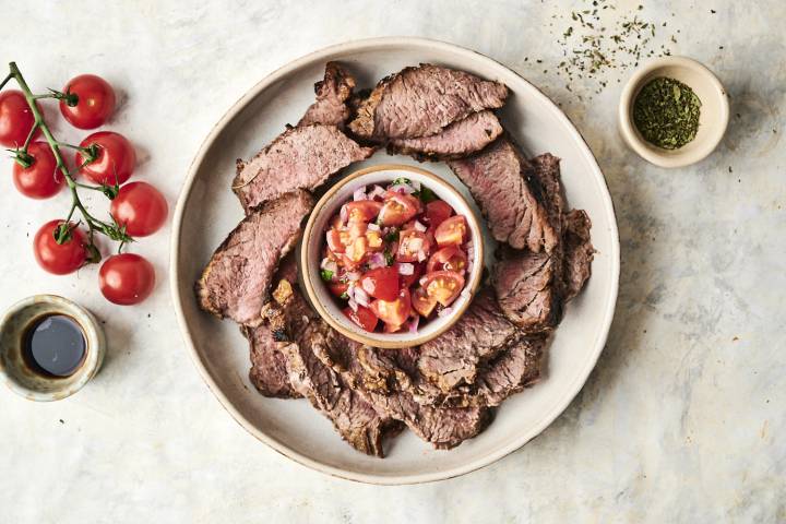 Spanish Flank Steak Platter of sliced beef with a bowl of fresh tomato salsa, surrounded by cherry tomatoes and spices on a light background.