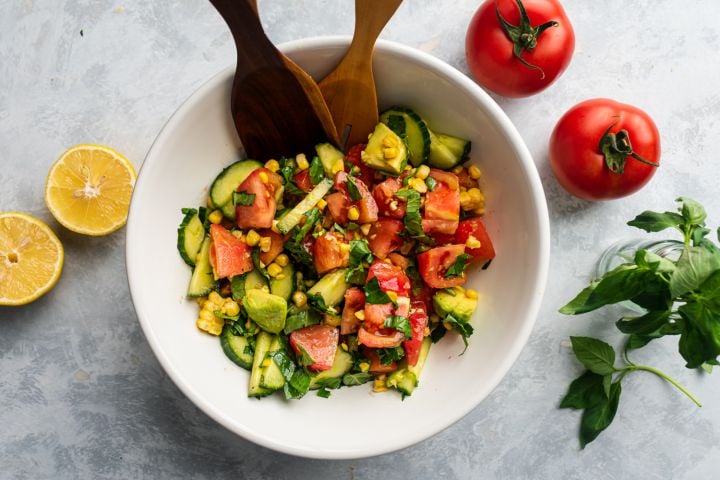 Tomato and Cucumber Salad with Corn Tomato cucumber salad with corn, avocado, and fresh basil in a bowl with wooden salad tongs.